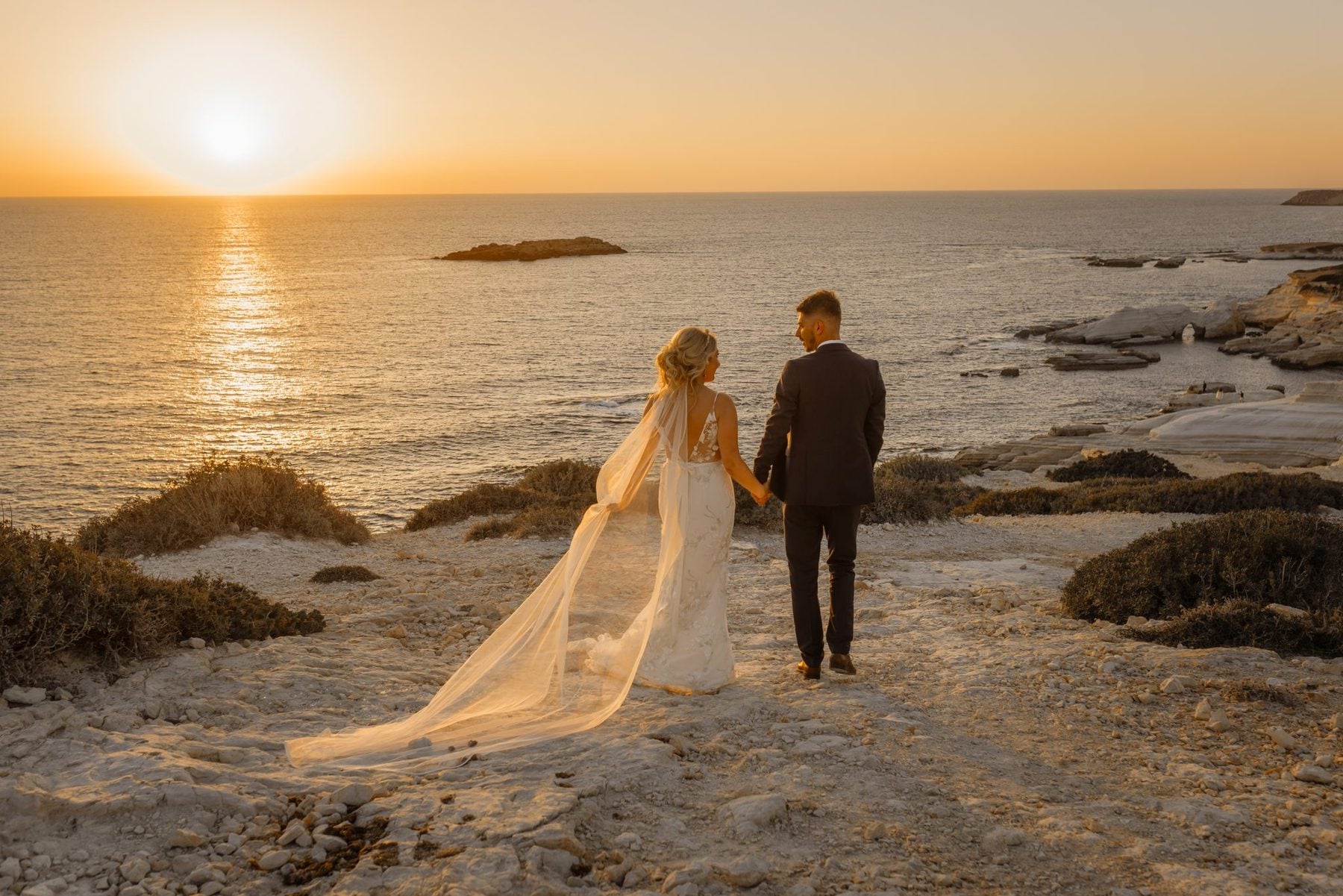 Bride and groom walking across the beach at Coral Residences towards the sea at sunset