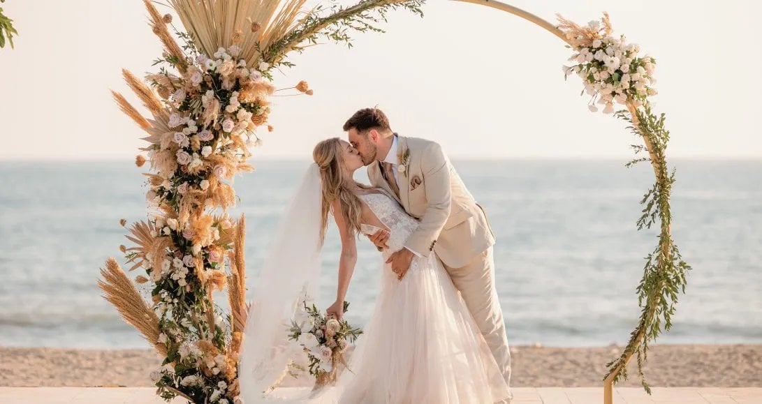 Bride and groom kissing beneath a floral arch on the beach at Coral Residences