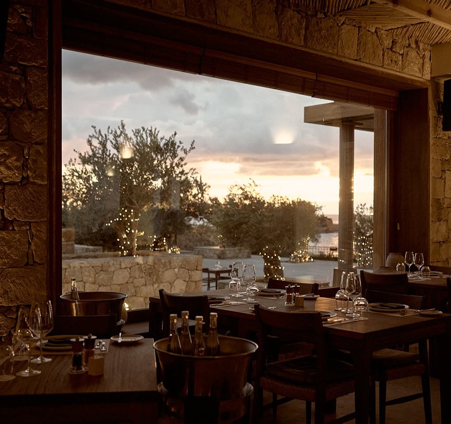 Set tables at PHOS restaurant with a view through large windows to olive trees and the sea at dusk